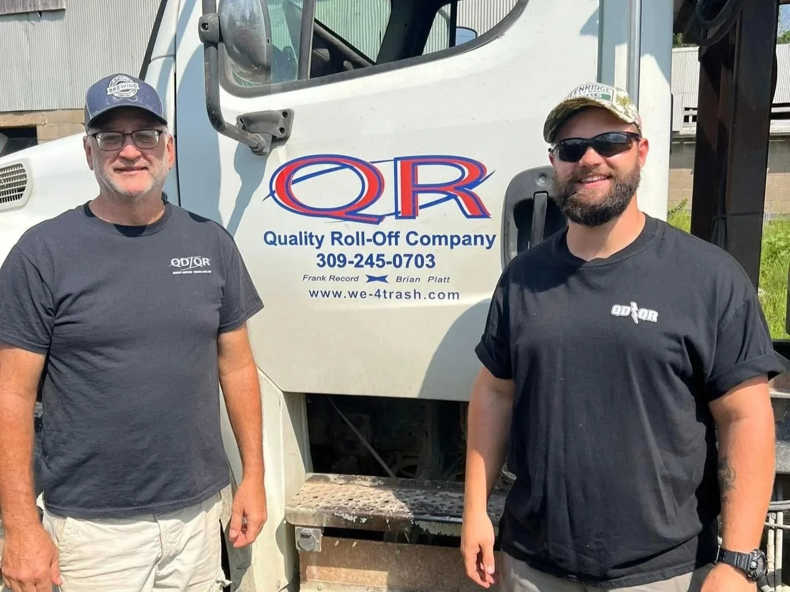 Two men standing outdoors in front of a white truck with a company logo and contact information. Both are wearing black T-shirts with the company logo, and the man on the right is wearing sunglasses and a cap.