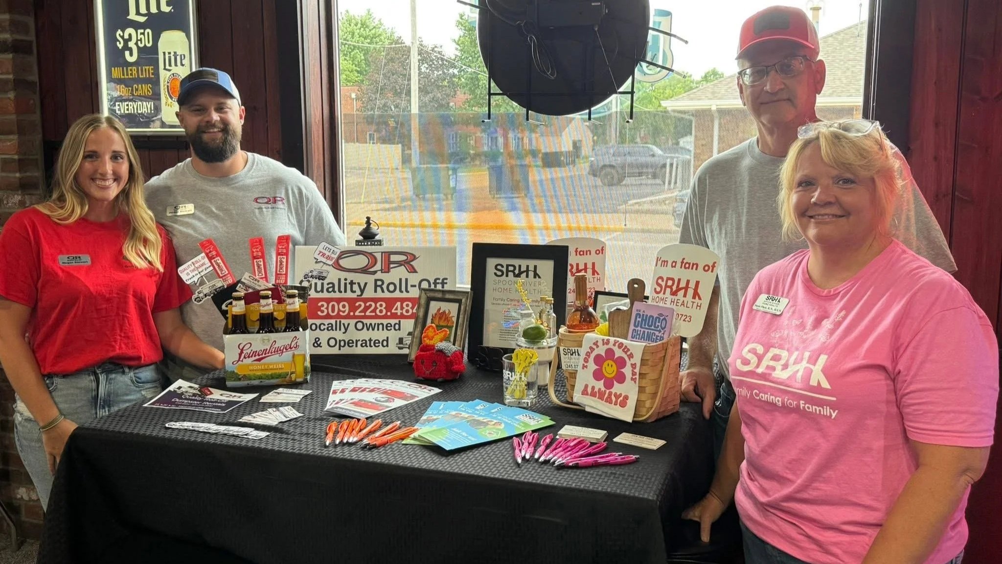 Group of five people standing behind a table with promotional items and signs, indoors near a window, with a parking lot outside.