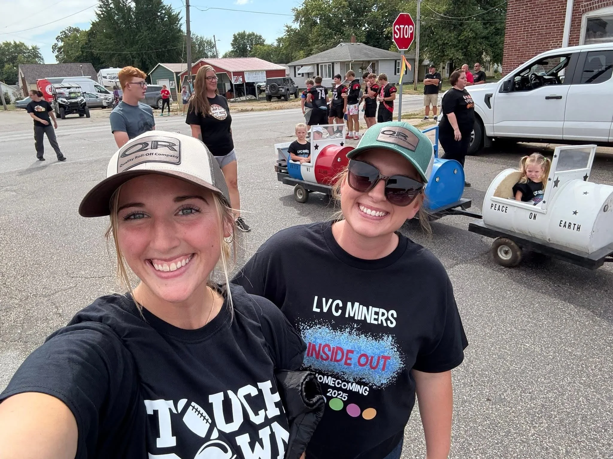 Two smiling women taking a selfie at a parade or community event, with people and decorated carts in the background, including a small cart labeled 'PEACE ON EARTH' and others designed to resemble vehicles.