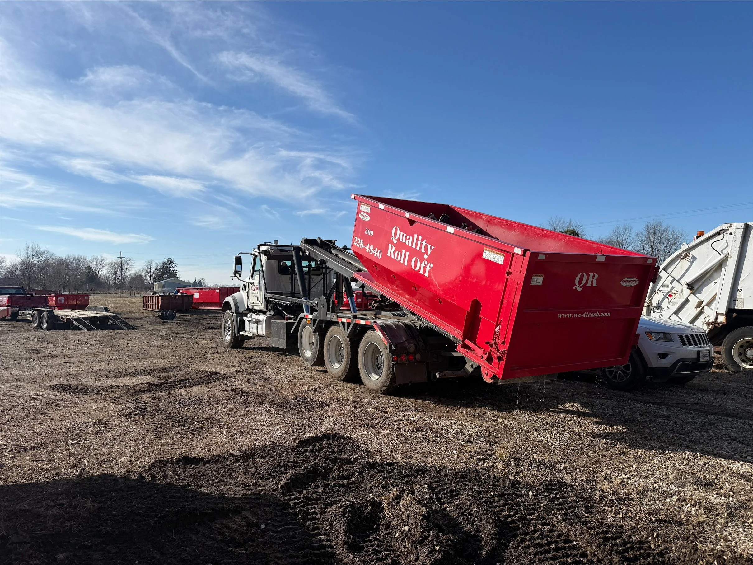 A dump truck with a red bed raised on a dirt lot, with several other trucks and trailers in the background under a blue sky with scattered clouds.