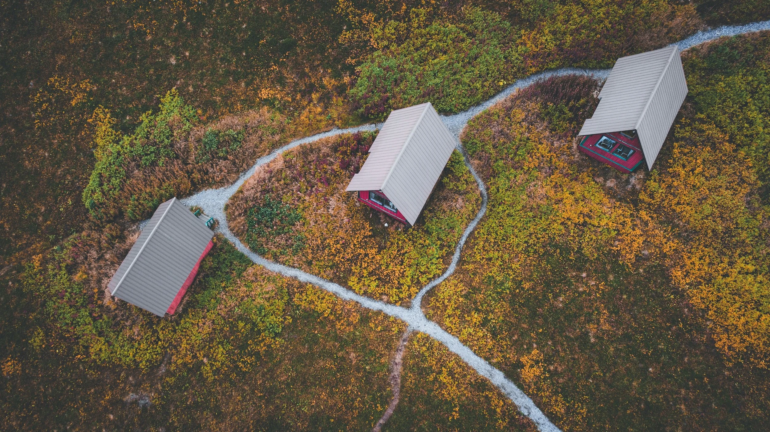An aerial view of three small red cabins with gray roofs in a forested area during autumn, with winding gravel paths connecting them.