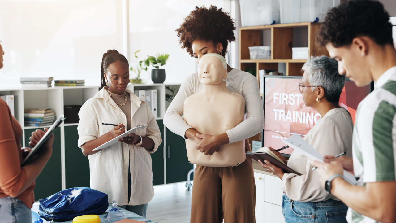 A woman holding a medical mannequin torso in a classroom or training setting with four other women, all taking notes, and a screen in the background reading 'First Training'.