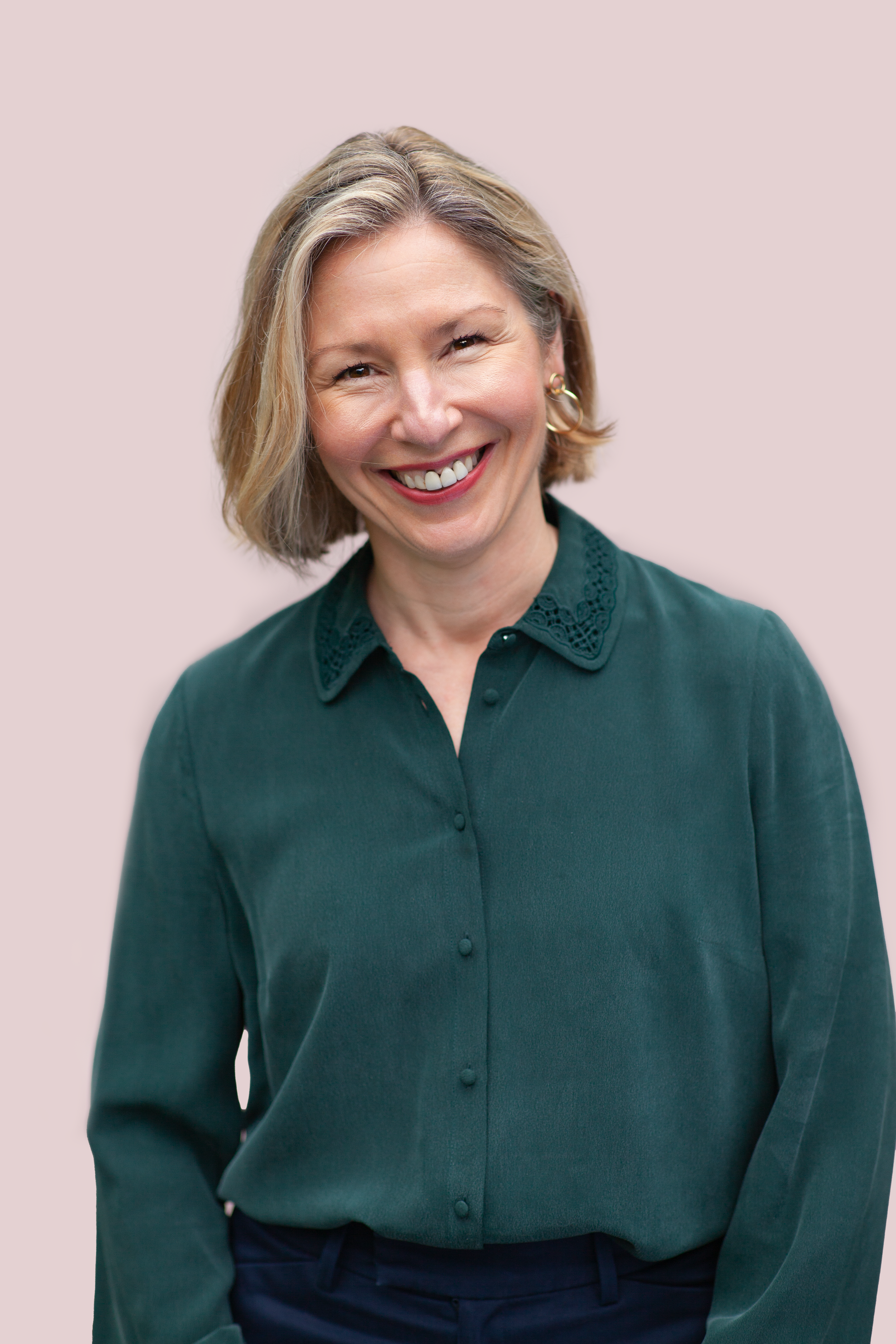 A smiling woman with short blonde hair, wearing a dark green button-up shirt with lace details on the collar, against a light pink background.