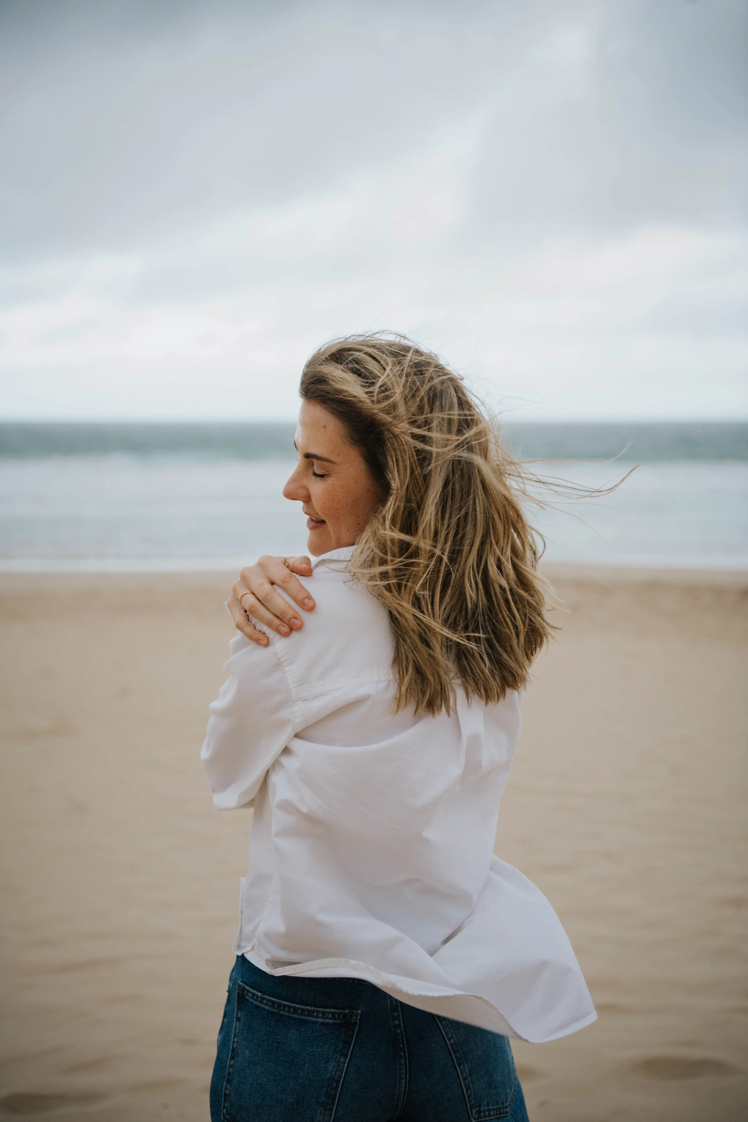 A woman with long, wavy hair, wearing a white shirt and blue jeans, standing on a beach with her eyes closed and a smile, embracing herself under an overcast sky.