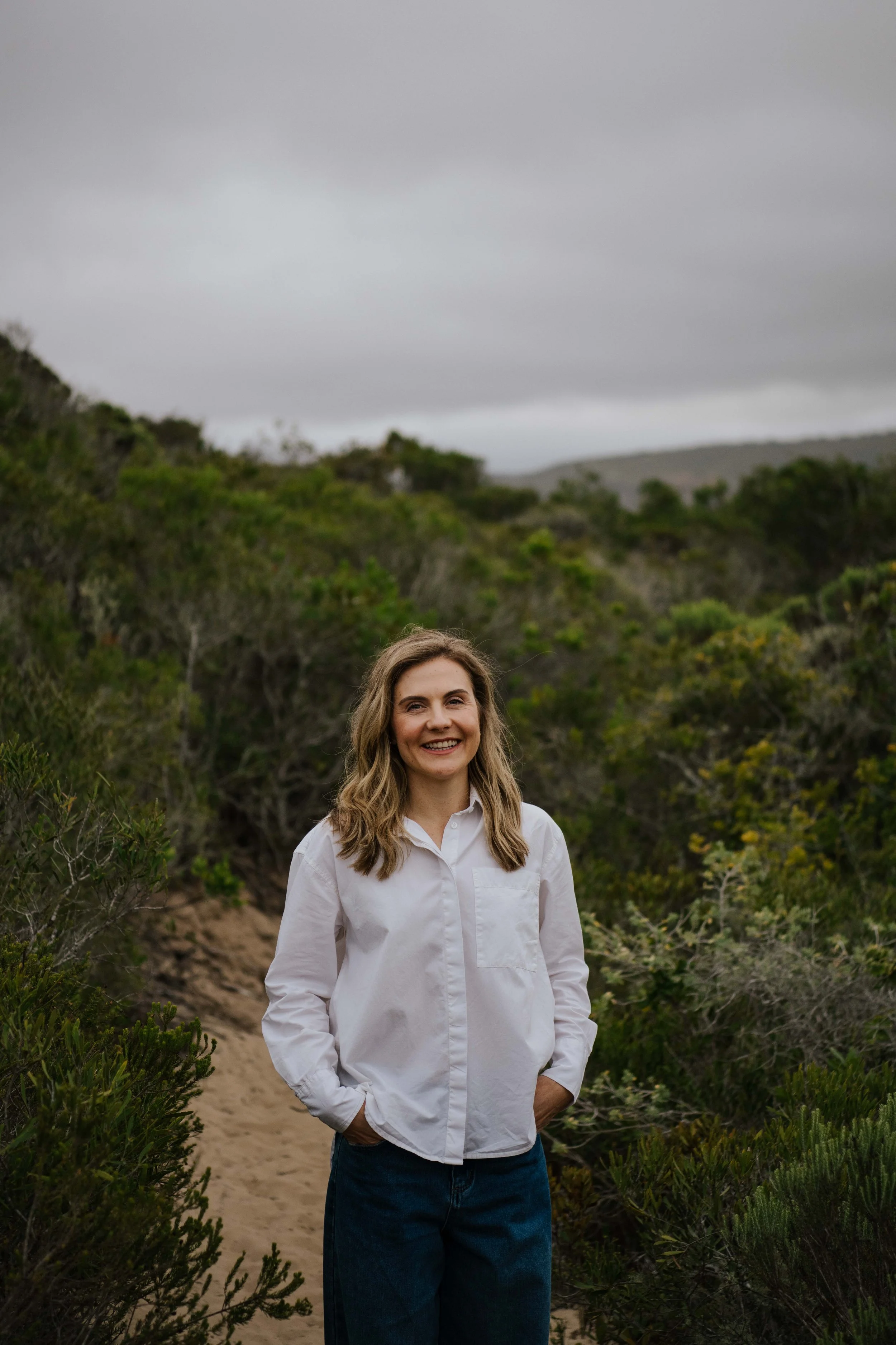 A smiling woman with blonde hair wearing a white shirt and blue jeans stands on a trail surrounded by green bushes and trees, under a cloudy sky.