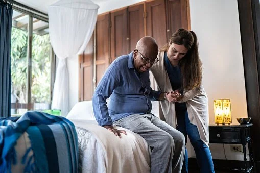 A woman assisting an elderly man with walking in a bedroom.