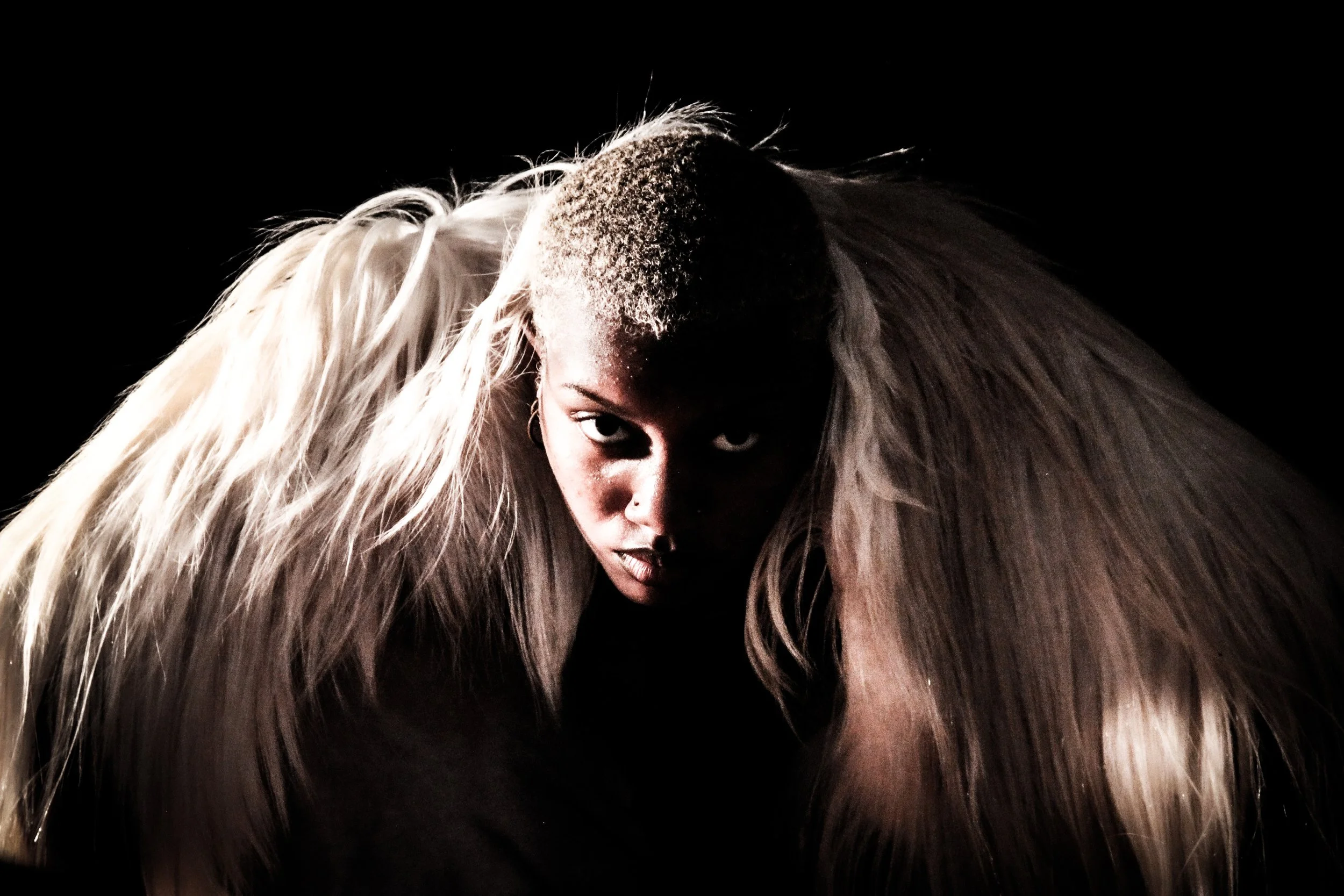 A person with platinum blonde hair in pigtails leaning forward, staring intensely at the camera against a black background.