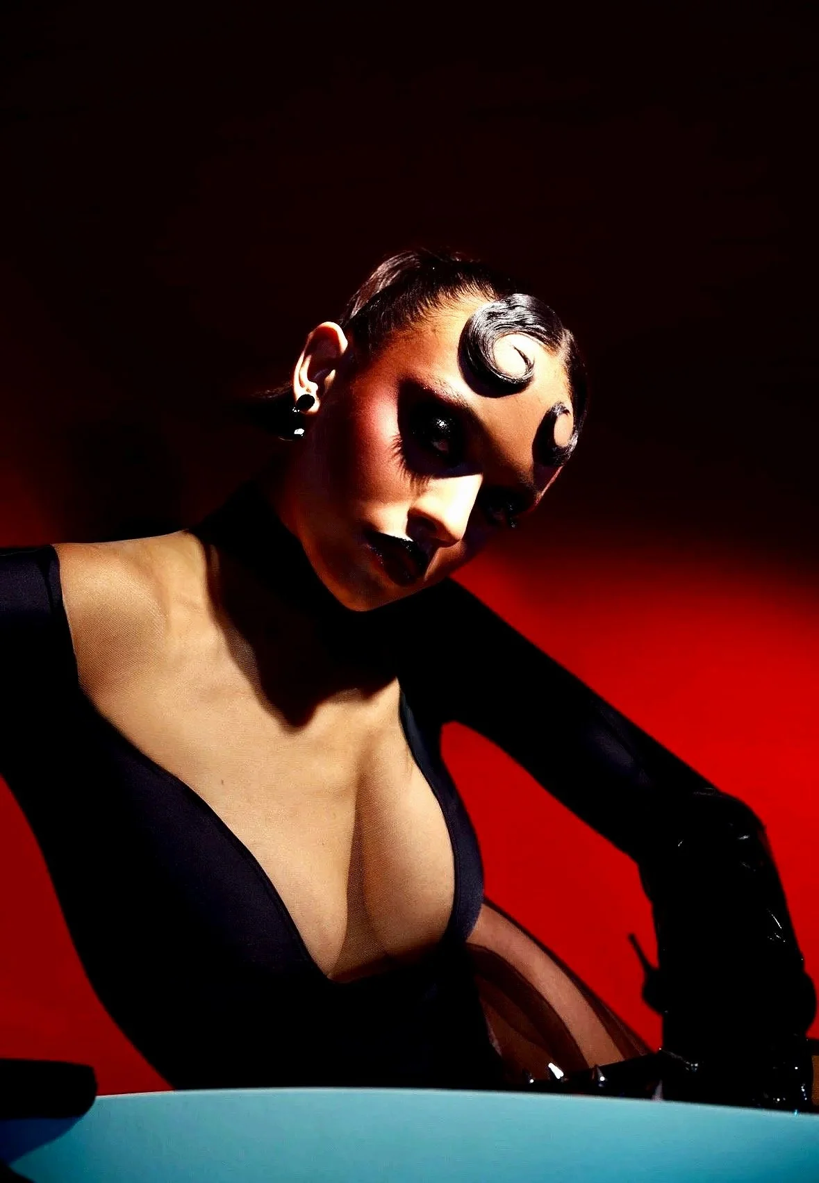 A woman with dark hair styled in vintage curls, dark eye makeup, and black lipstick, wearing a black dress with sheer sleeves, posing against a red background.