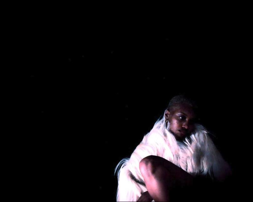 A person with platinum blonde hair in a white fur coat sitting against a black background.