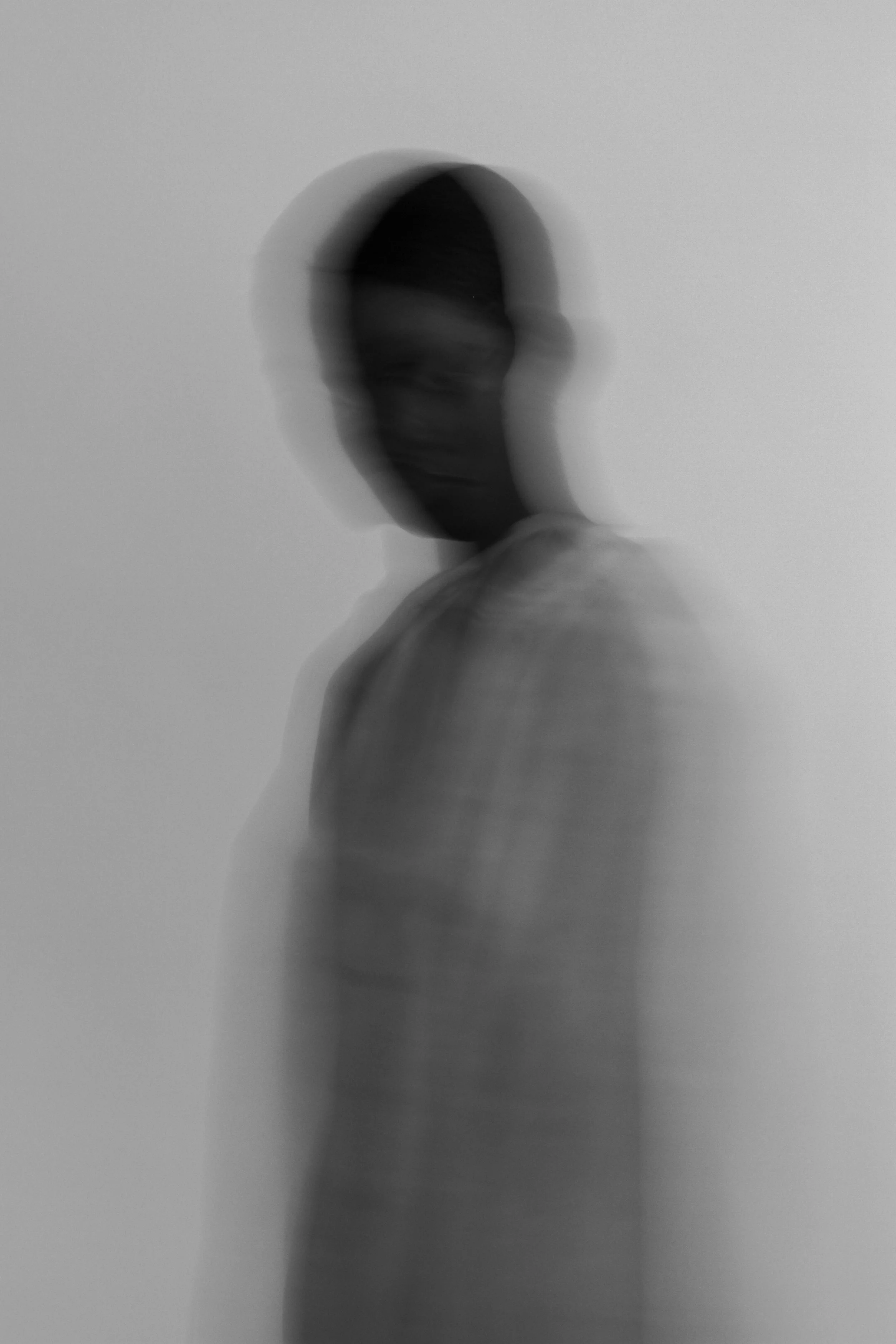 A black and white blurred photo of a person standing against a plain background, with a motion trail effect.