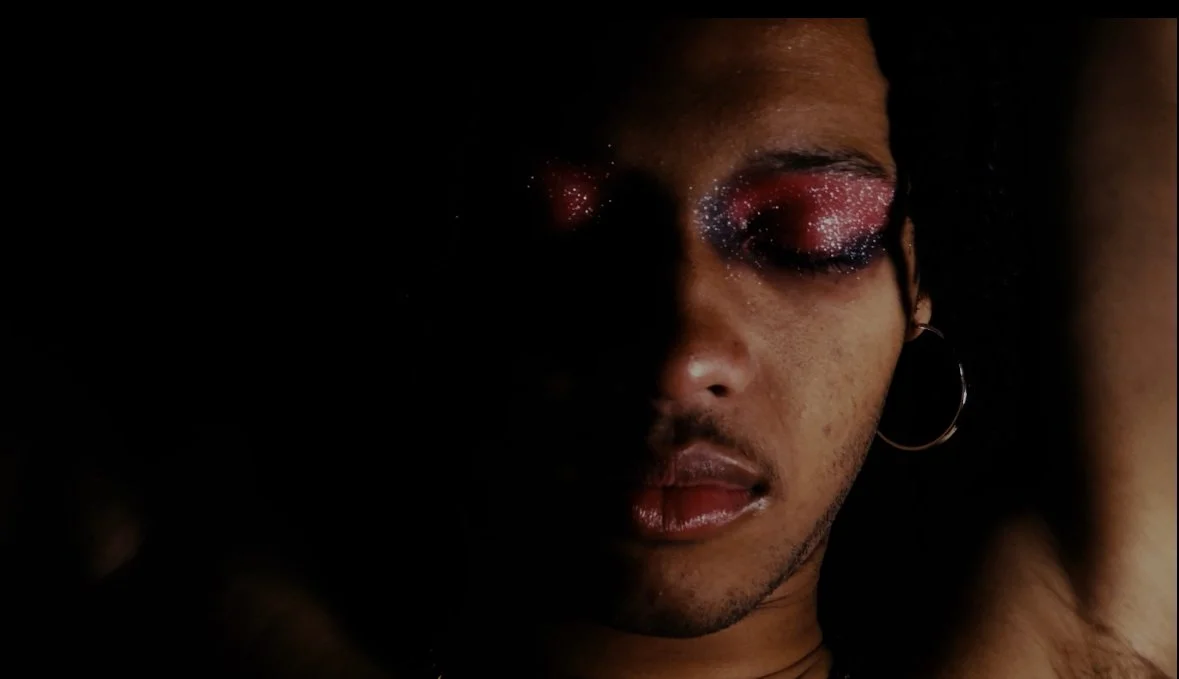 A close-up of a person with glittery, colorful eye makeup and hoop earrings, with a dark background.