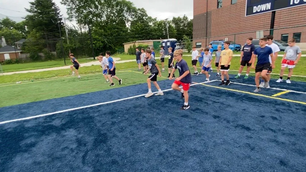 Group of boys participating in a running drill on a sports field with blue and green turf, near a brick building with a sign that says 'DOWN'.