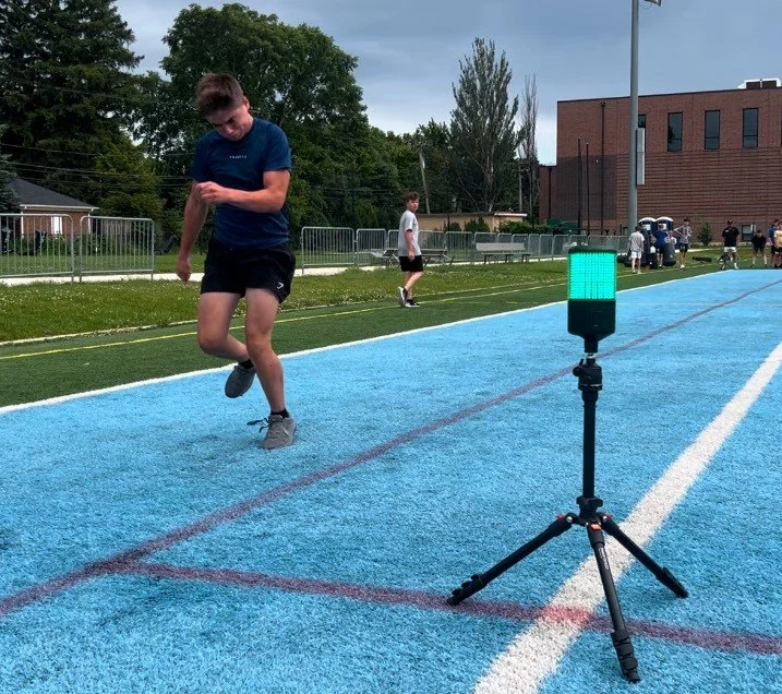 Young male athlete running on a blue track, with a timing device on a tripod in the foreground, during daytime.
