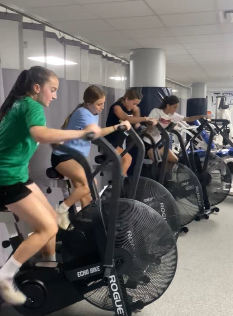 Four girls using stationary exercise bikes in a gym, focused on their workout.