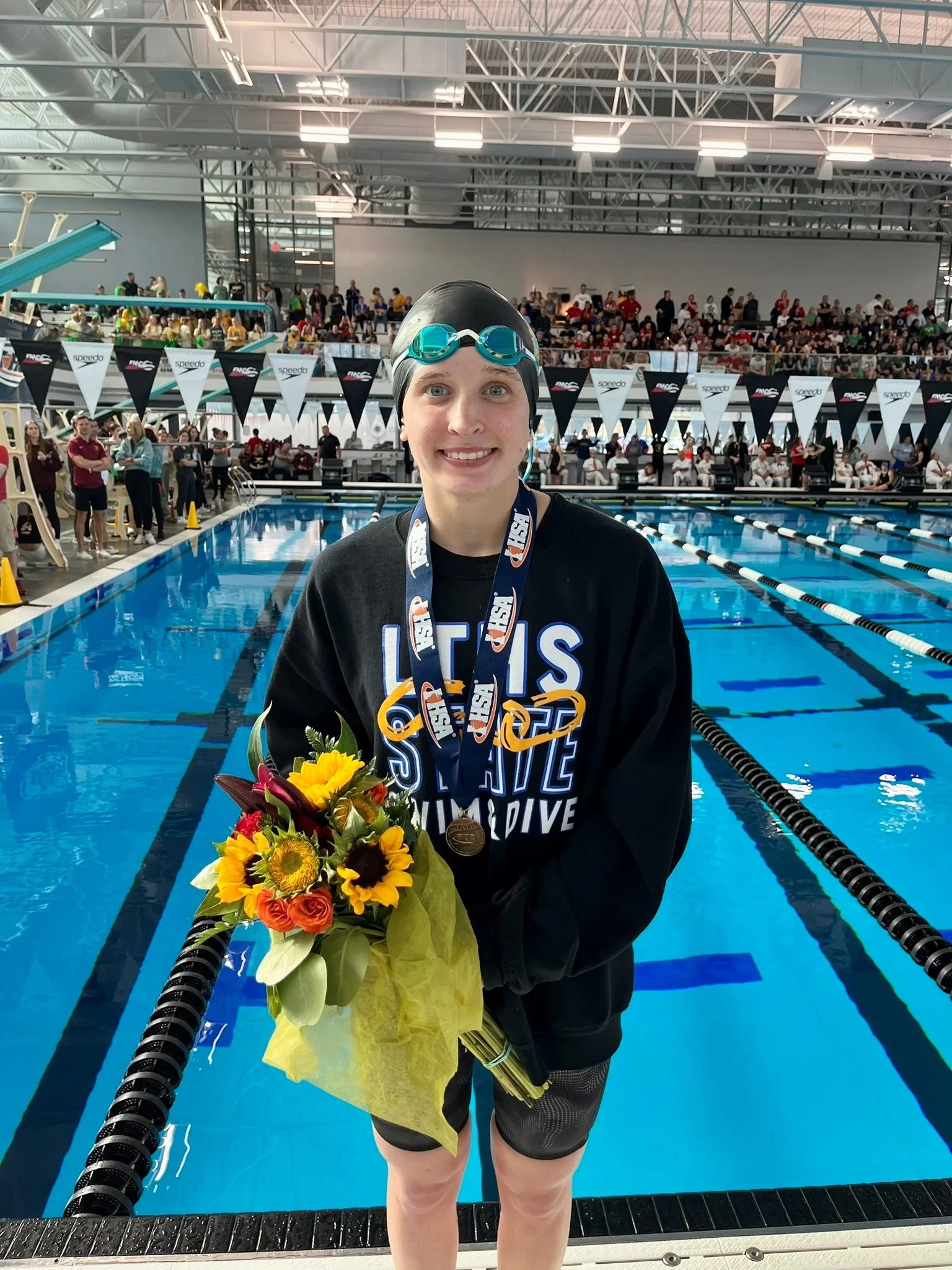 Swimmer with goggles and swim cap, holding a bouquet of flowers and wearing medals, standing by an indoor swimming pool during a competition.