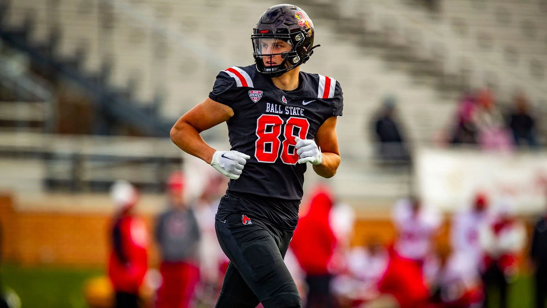 A football player wearing a black uniform with red accents, helmet, and gloves is running on the field.