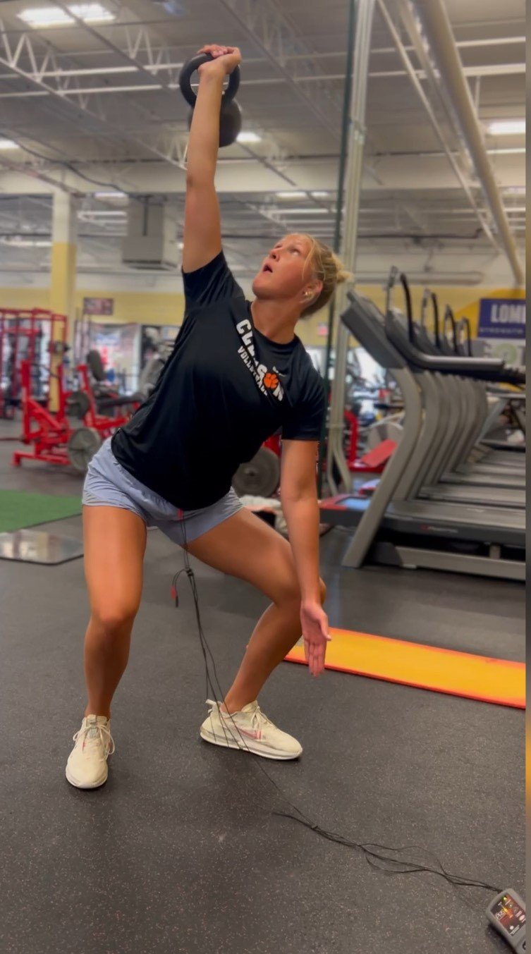 A woman in workout attire lifting a kettlebell in a gym, with gym equipment and treadmills in the background.