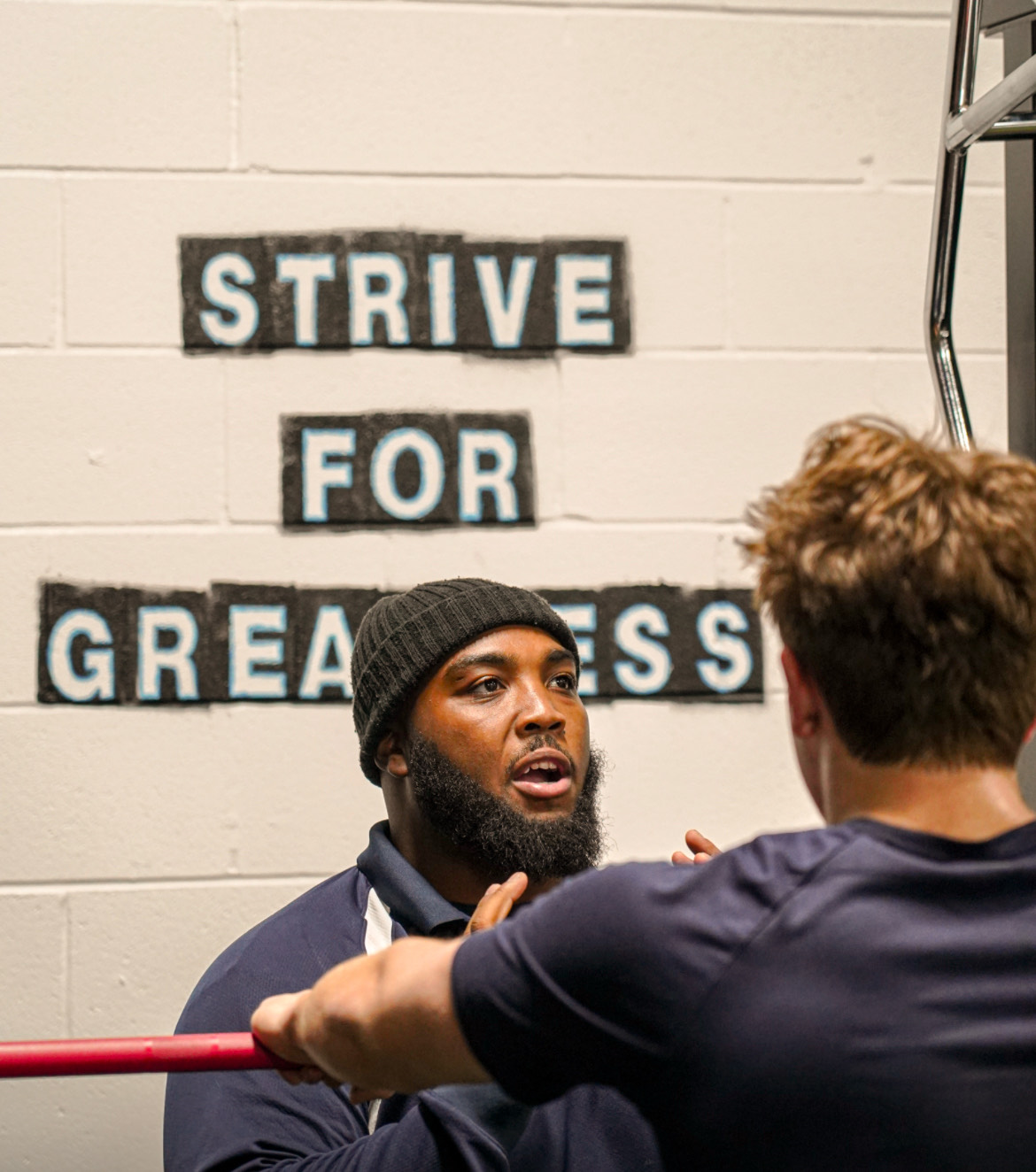 Two men in a gym, one holding a red barbell, engaged in a conversation. In the background, a wall with a motivational quote that reads 'STRIVE FOR GREATNESS'.