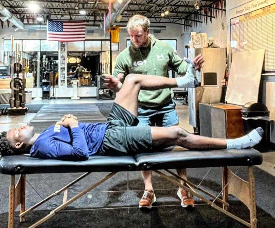 A person lying on a massage table with their leg being stretched or manipulated by a professional therapist in a gym or physical therapy facility.