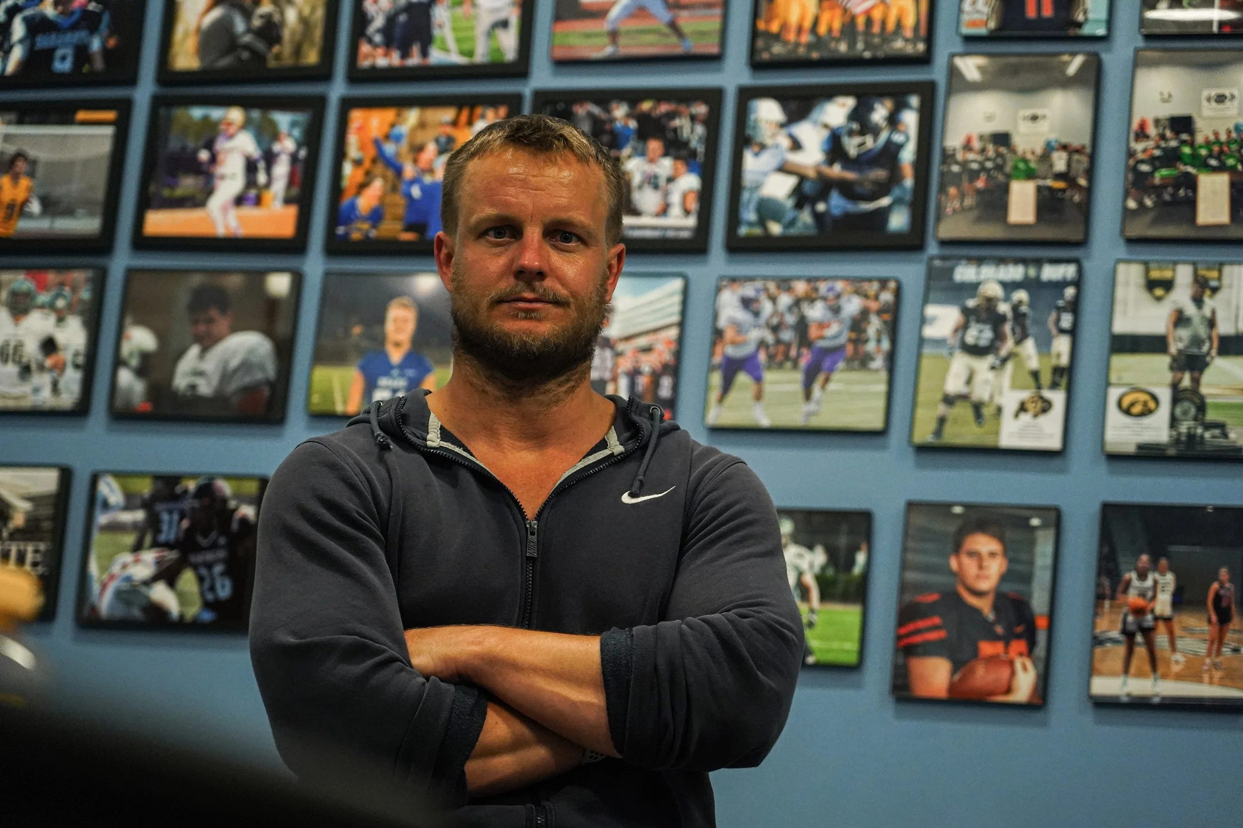 A man with a beard and short hair wearing a dark Nike zip-up hoodie, standing with arms crossed in front of a wall decorated with framed sports photographs.