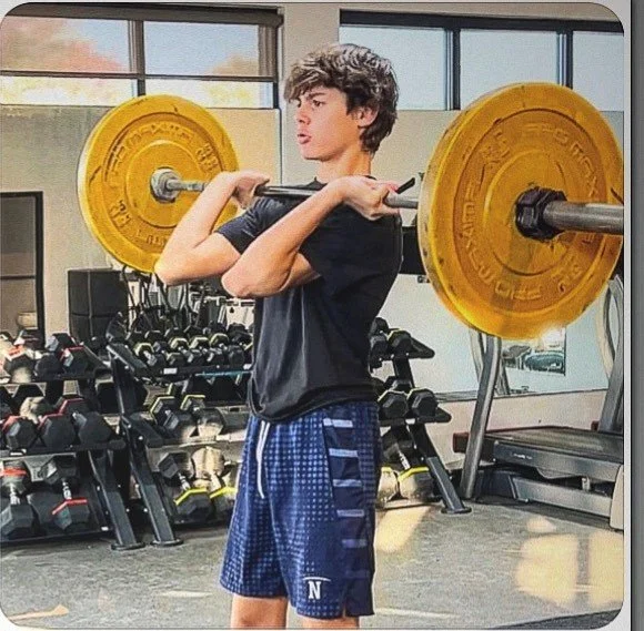 A teenage boy lifting a barbell with large yellow weight plates on each end in a gym.