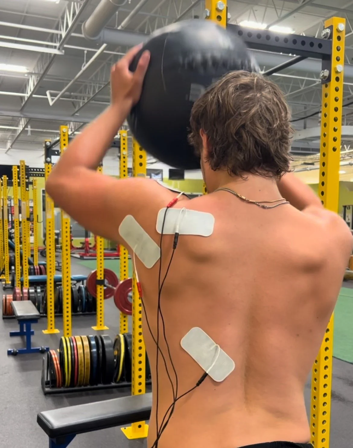 A man with a shirtless back lifting a black medicine ball in a gym, with electrodes and bandages attached to his back and shoulder.