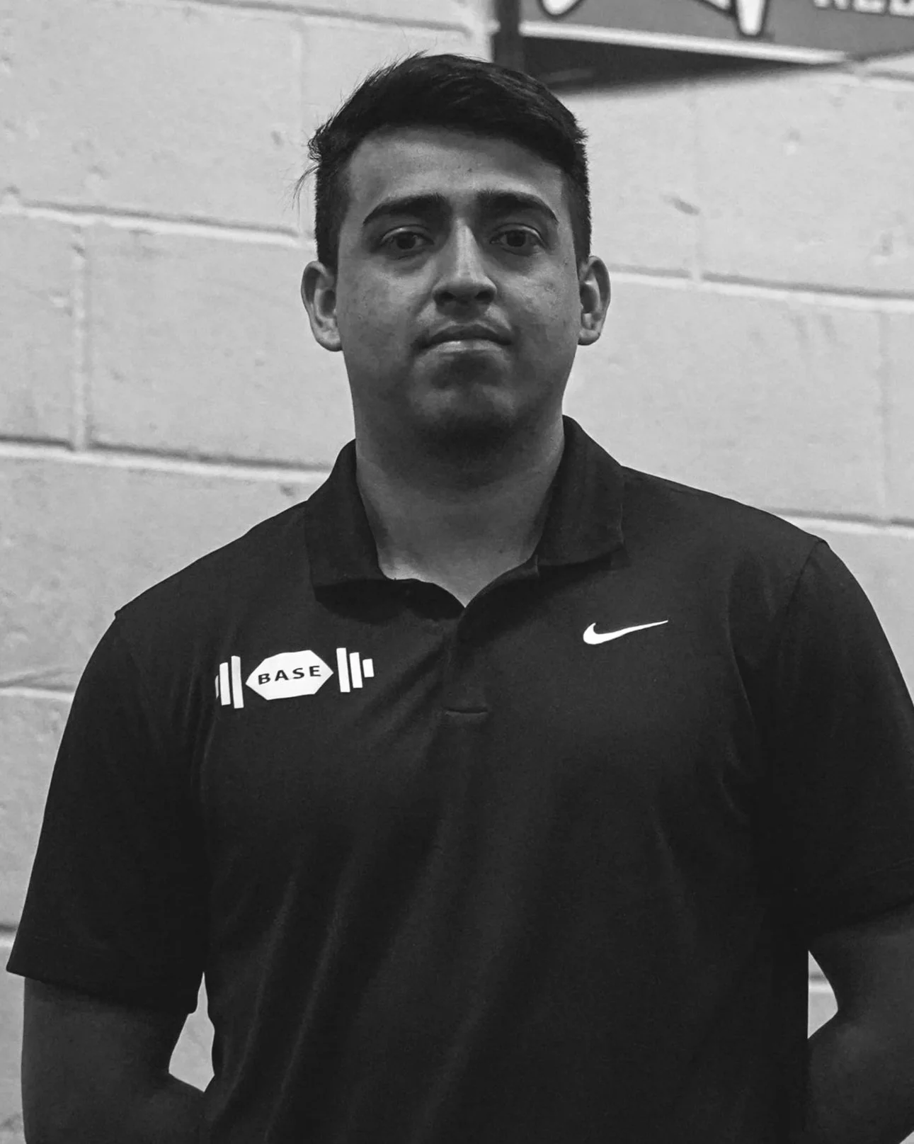 A young man with short dark hair and a serious expression, wearing a black Nike polo shirt with a 'BASE' logo and a white Nike swoosh, standing against a brick wall in black and white - Julian Hernandez Personal Trainer