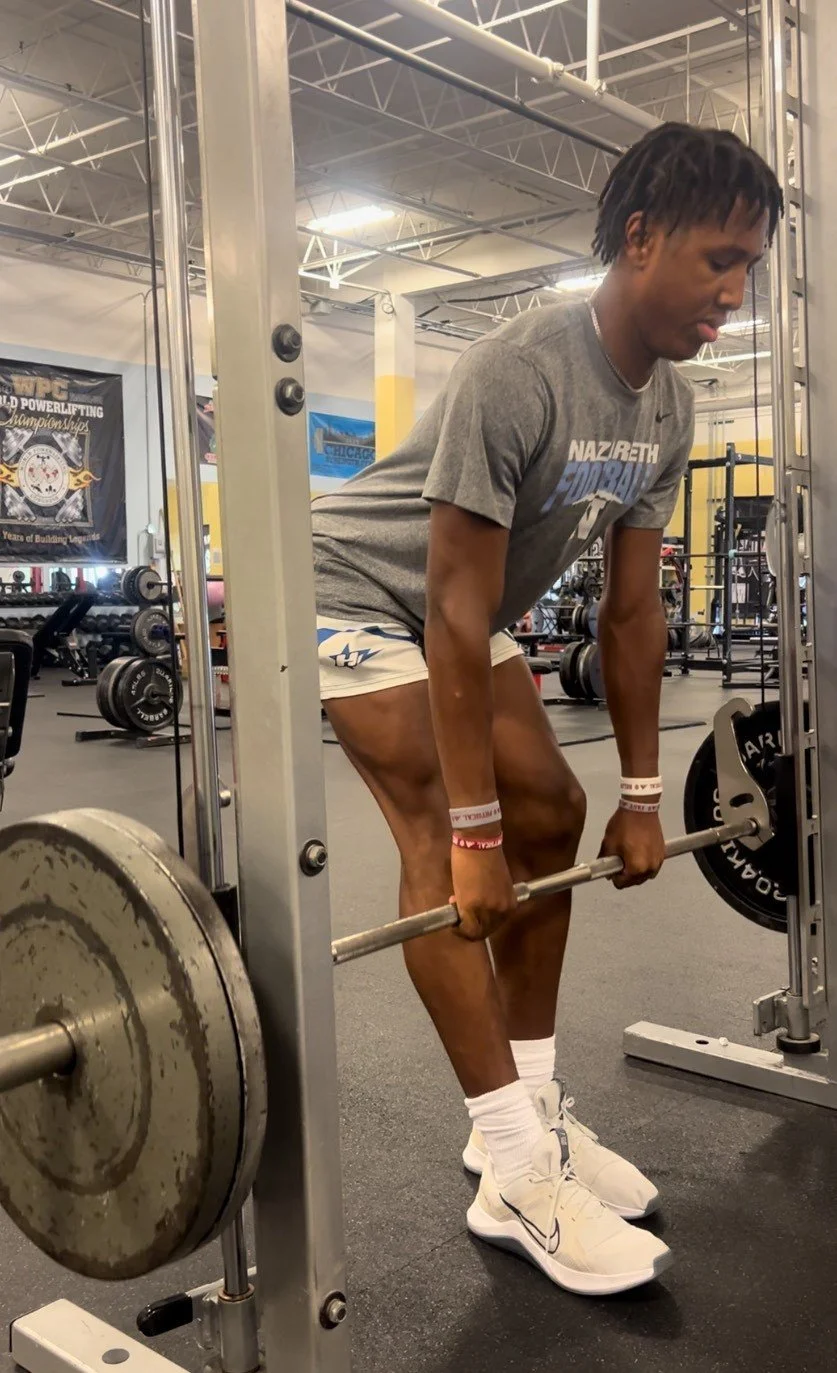 A young man lifting a barbell with weights in a gym, wearing a gray t-shirt, white shorts, and white Nike sneakers.