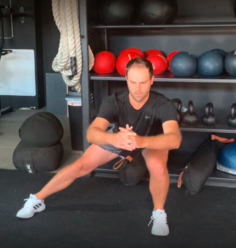 Man performing a side squat exercise in a gym, with black shelves holding red and blue medicine balls behind him.