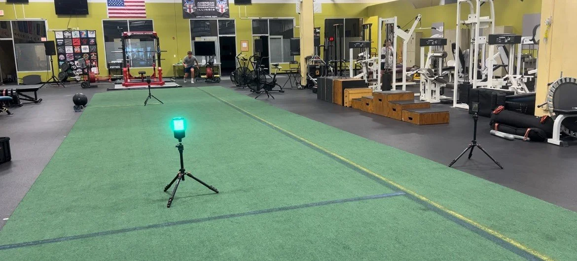 Equipment and exercise area inside a gym with a green laser level on a tripod, weightlifting machines, benches, and people working out.