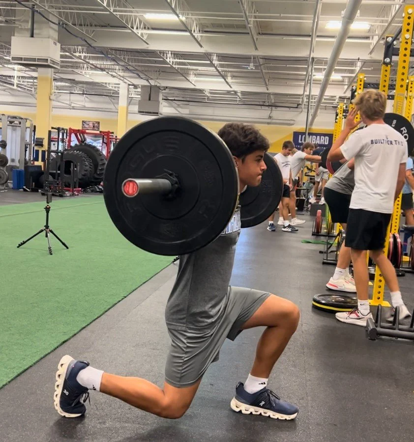 Young man performing a lunge with a barbell across his upper back in a gym.