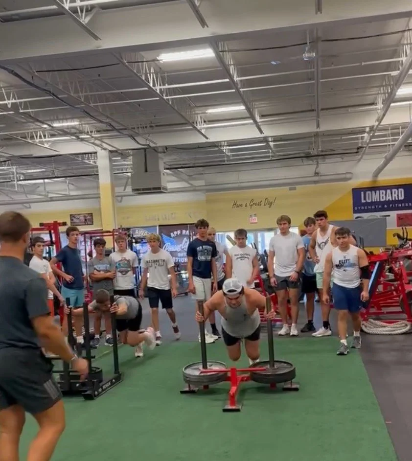 A group of young men at a gym watching a person push a weighted sled across a green turf.