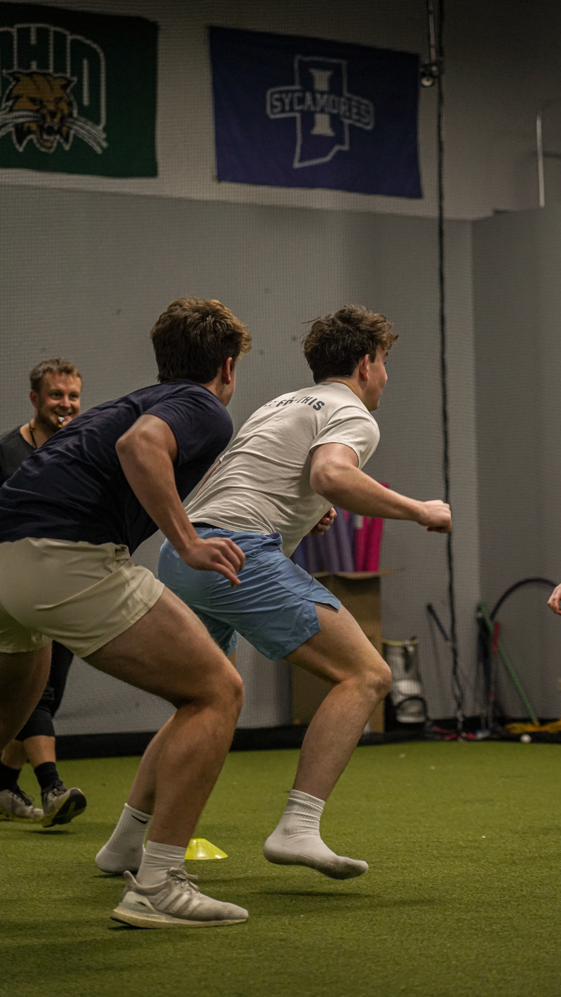 Three young men participating in a sprint race training indoors, with two in front and one smiling behind them, on a green turf with flags hanging on the wall.