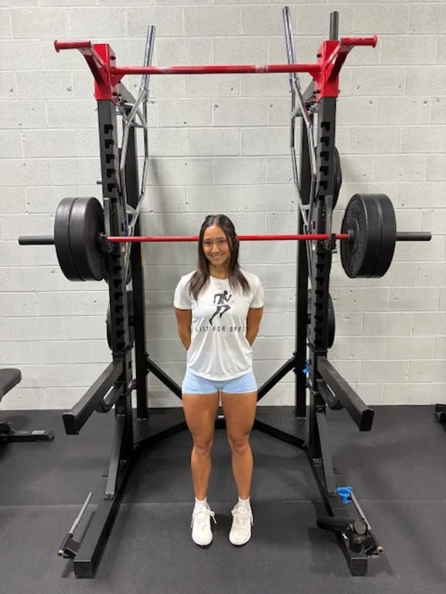 A woman standing in front of a large squat rack with a barbell loaded with weights. The woman is smiling and wearing a white T-shirt, blue shorts, and white sneakers. The squat rack is in a gym with a gray brick wall background.