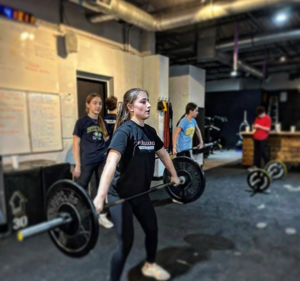 A young woman lifts weights in a gym with several people in the background, including a girl in a black shirt, a person in a light blue shirt, and another person in a red shirt near a wooden counter.