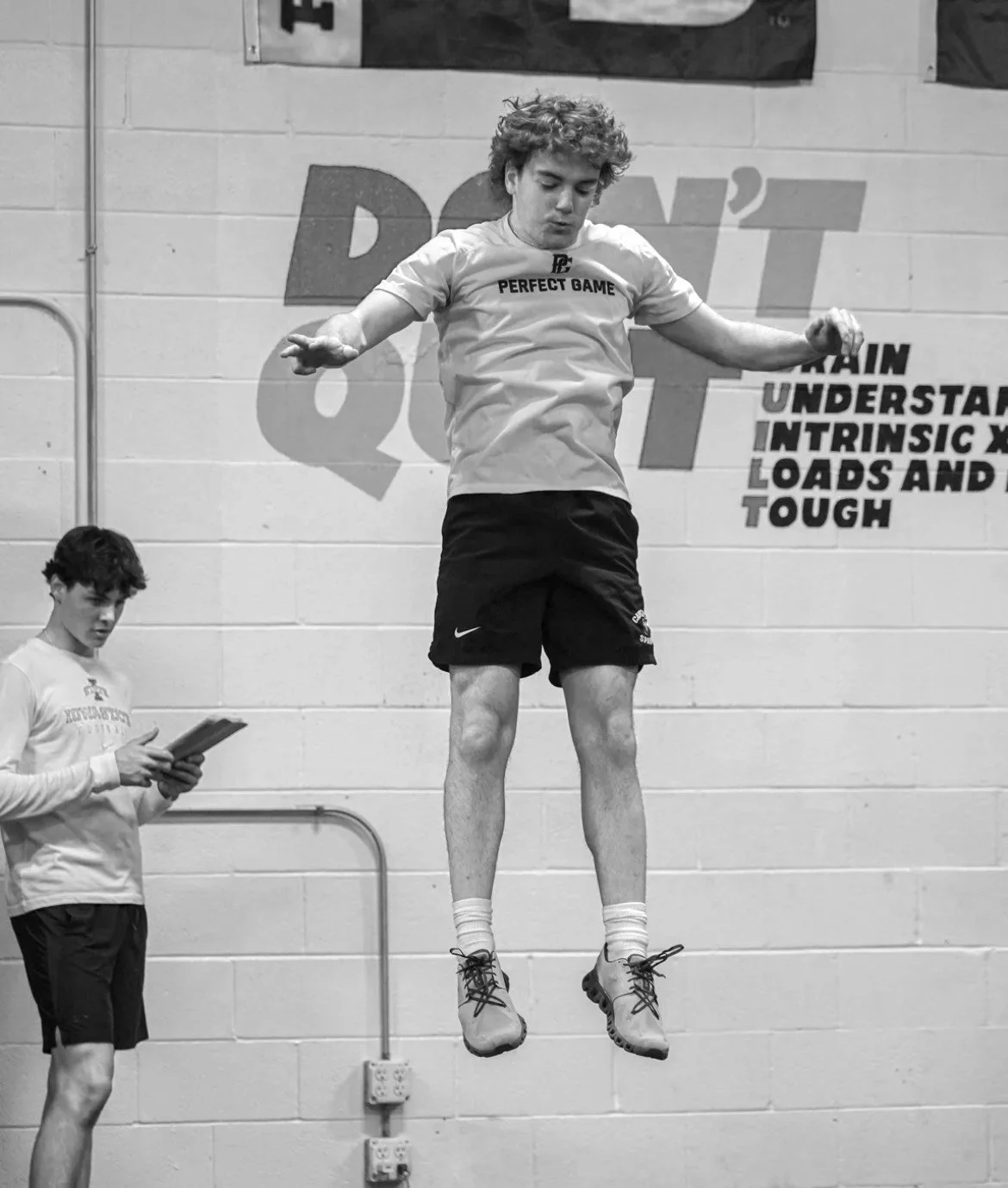 A young male athlete in a gym is captured mid-air during a jump training exercise, wearing a T-shirt and shorts, with another person holding a clipboard nearby. The background features motivational text on the wall.