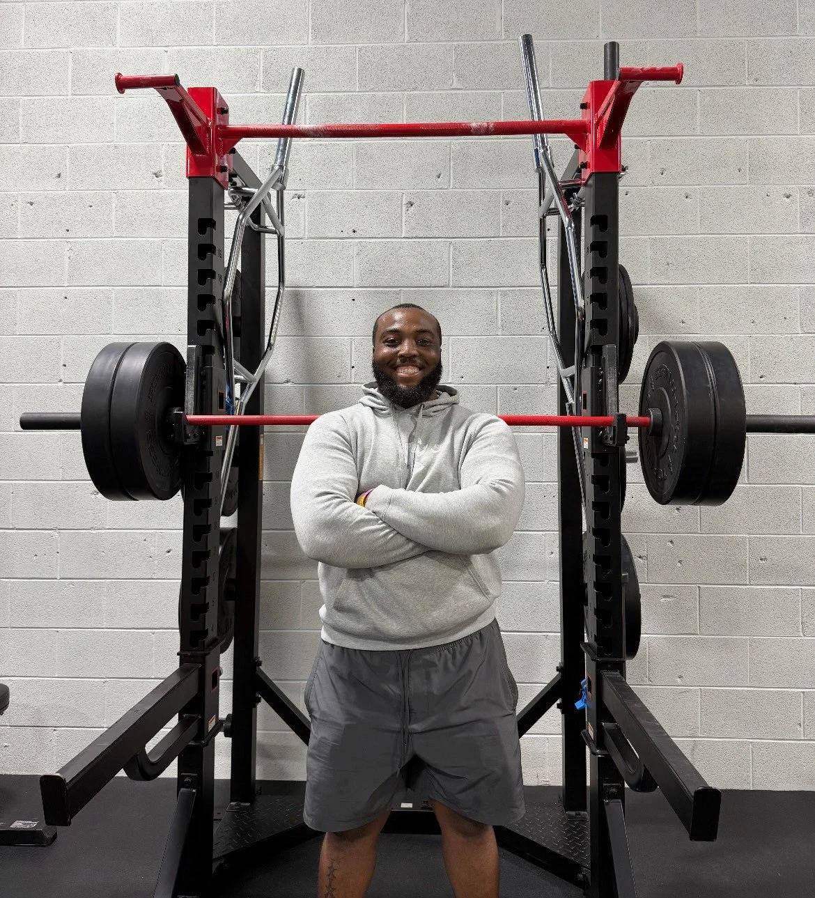 A smiling man standing in front of a weightlifting rack with arms crossed, in a gym with a concrete block wall.