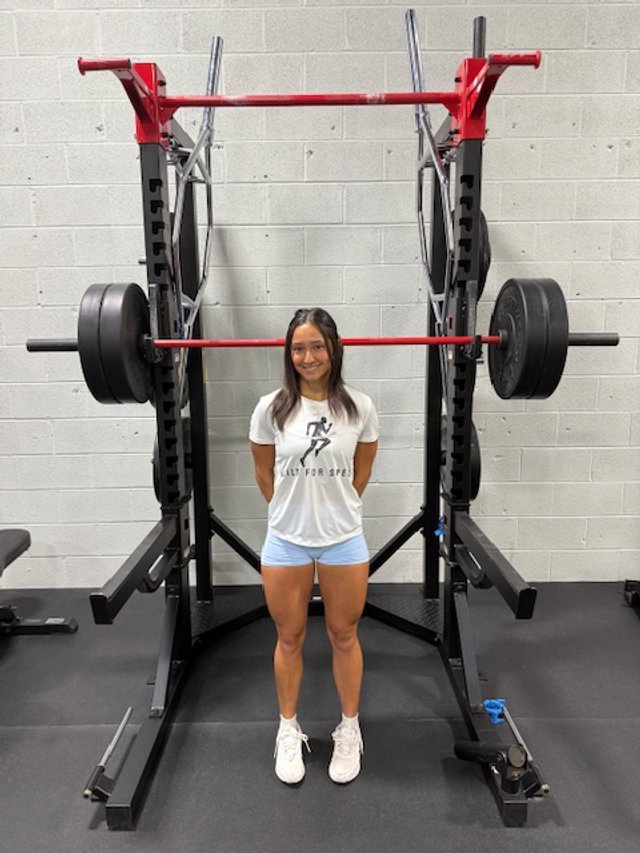 A woman standing in front of a large squat rack with loaded weights in a gym.