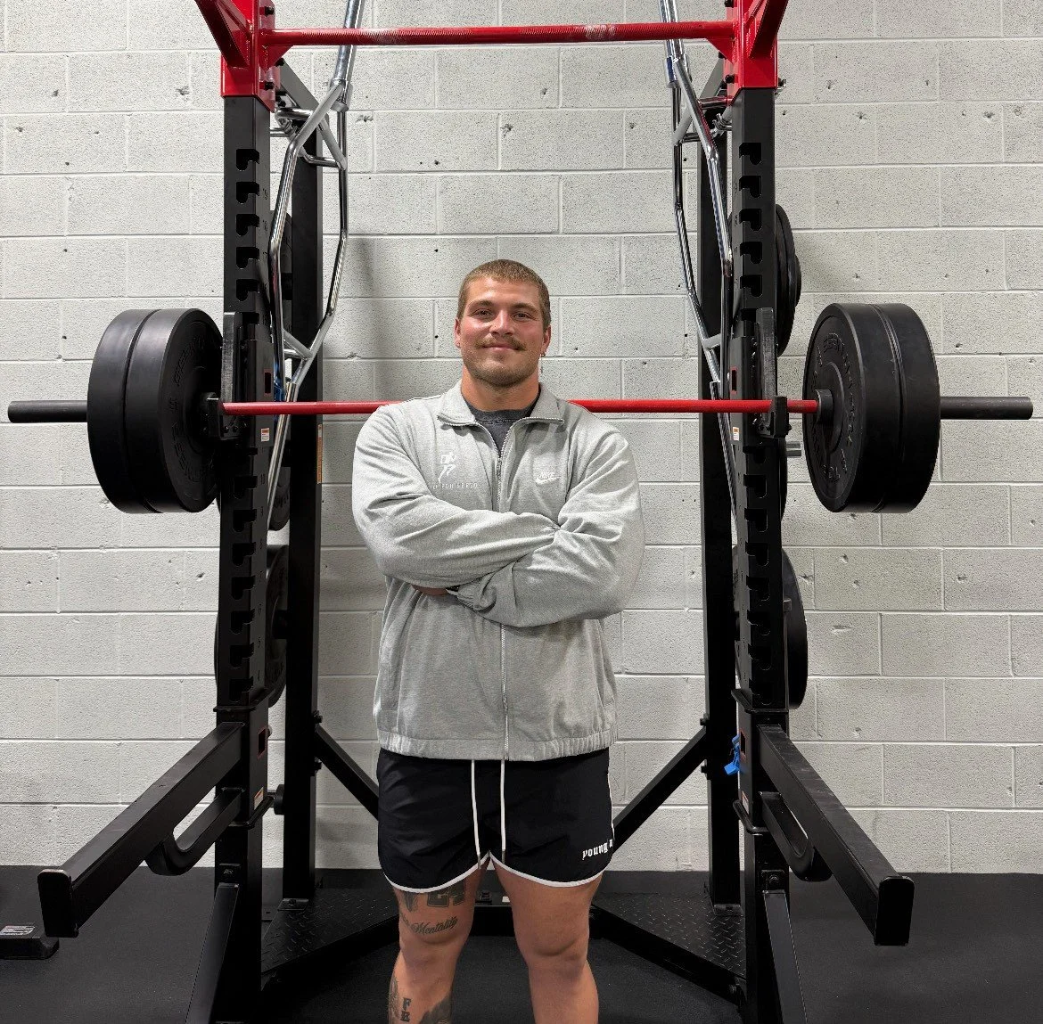 A man standing with arms crossed in front of a squat rack with weights in a gym.