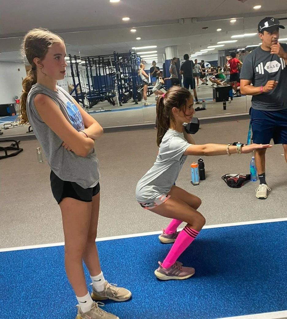 Two young girls in workout attire at the gym, one demonstrating a squat while the other observes, with gym equipment and people exercising in the background.