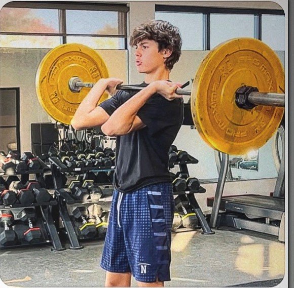 A young man in a gym performing a shoulder lift with a barbell loaded with yellow weight plates.