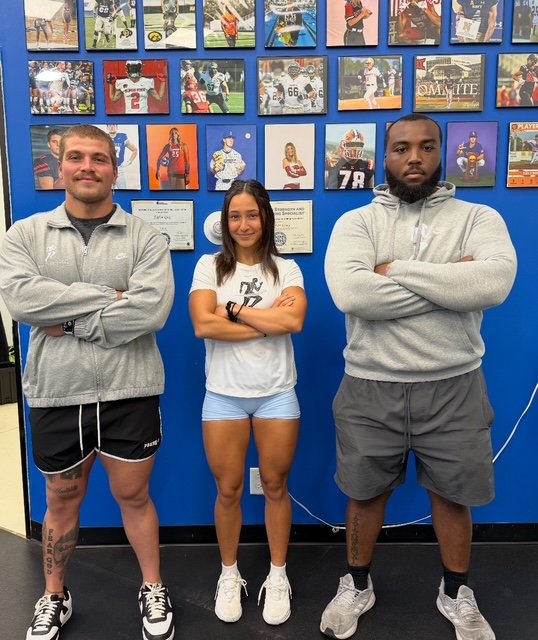 Three young athletes standing with arms crossed in front of a blue wall decorated with framed sports photos and memorabilia.
