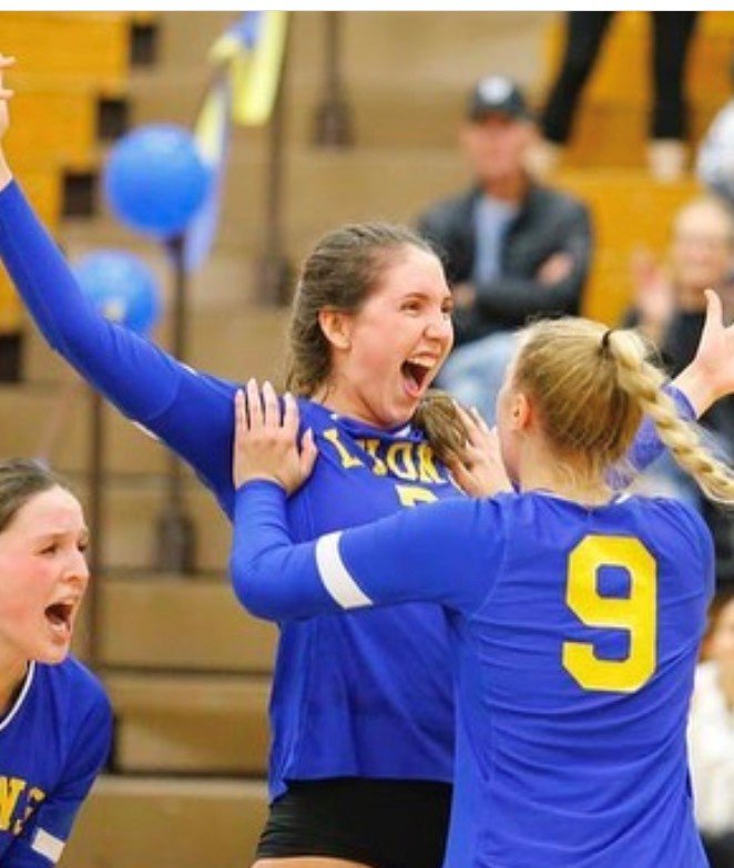 Three female volleyball players in blue jerseys celebrating on the court, with one girl in the center jumping and smiling widely, with teammates supporting her.