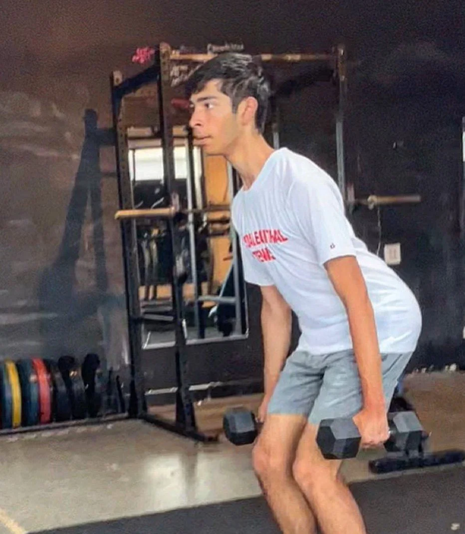 A young man with dark hair wearing a white shirt and gray shorts is exercising in a gym. He is holding a dumbbell in each hand and appears to be in a bent-over position for a workout.