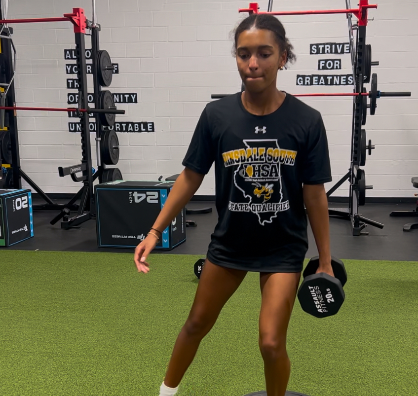Young woman working out at gym, lifting dumbbell, wearing black shirt with sports logo and shorts, gym equipment in background.