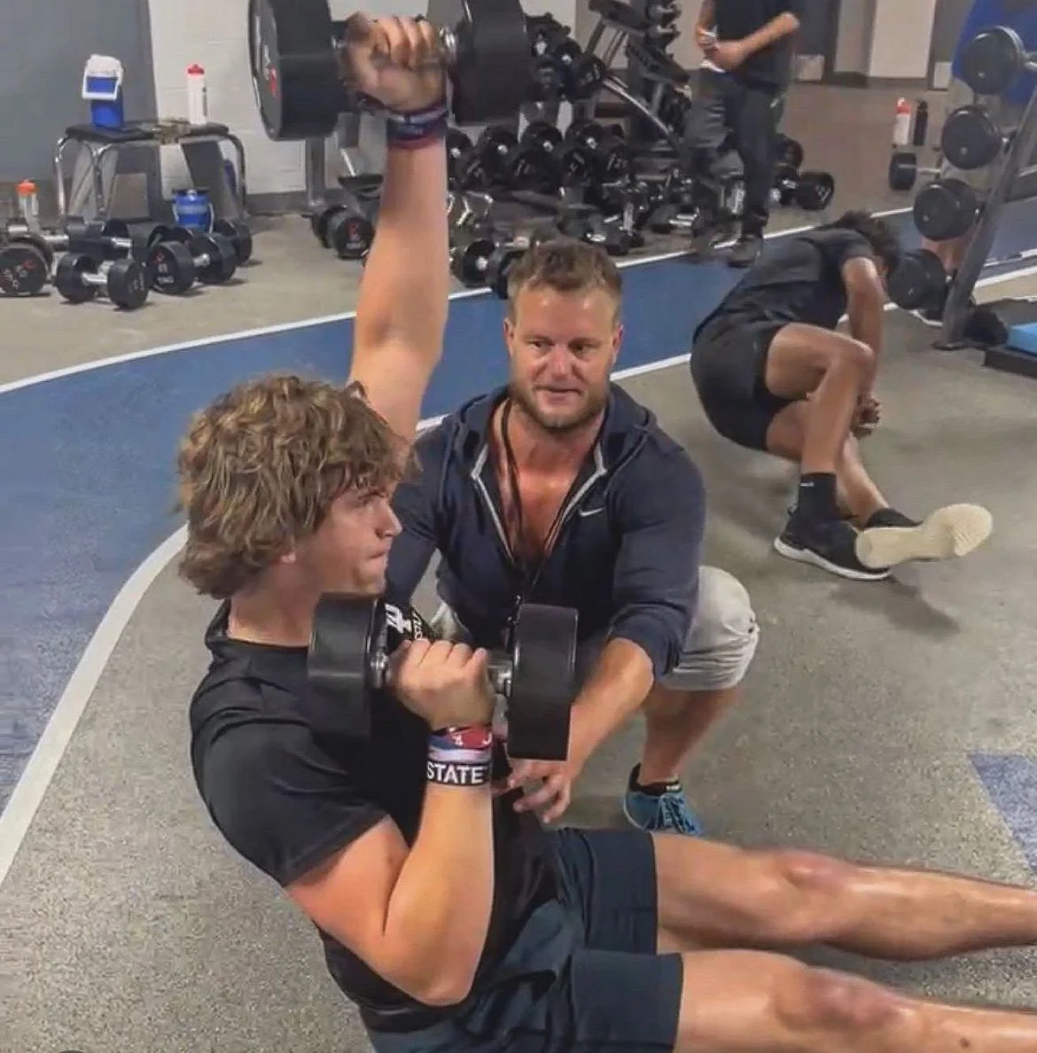 Two men are in a gym one is sitting on the floor lifting a dumbbell while a man kneeling behind him spots him. In the background, another person is doing an exercise on the floor and gym equipment is visible.