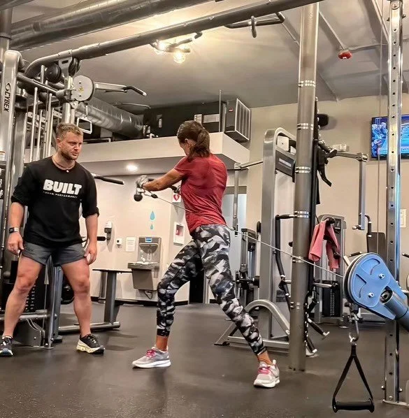 A woman in a red shirt and camouflage leggings is doing kettlebell swings at the gym, while a man in a black shirt and shorts supervises.