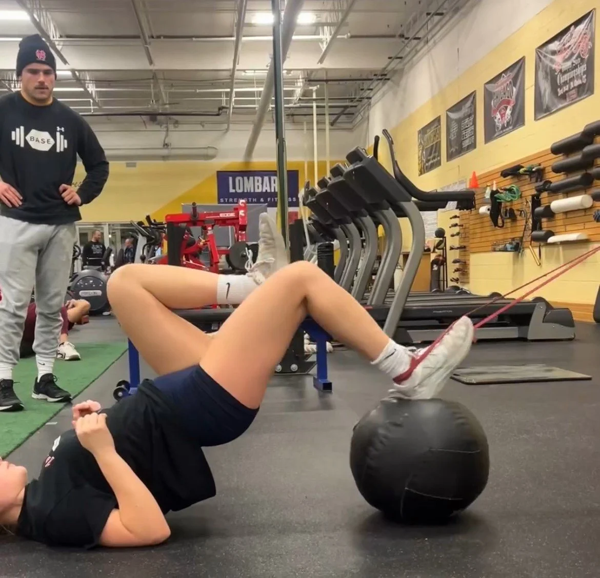 A woman performing a glute bridge exercise with her feet elevated on a black exercise ball in a gym, while a man and another person are standing nearby observing.