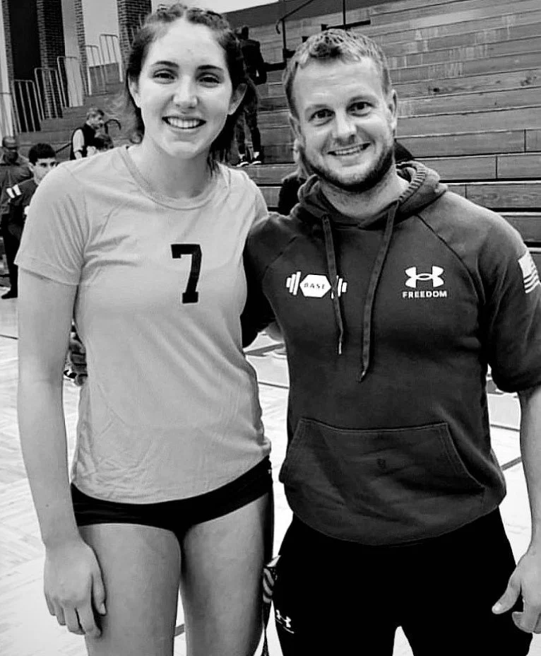 A young woman and a man smiling for a photo in a sports setting, with the woman wearing a volleyball uniform and the man in athletic hoodie and shorts.