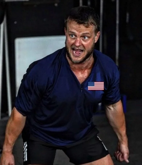Man wearing a blue shirt with a small U.S. flag patch, appearing to be exhausted or exerted, in a gym or fitness setting.